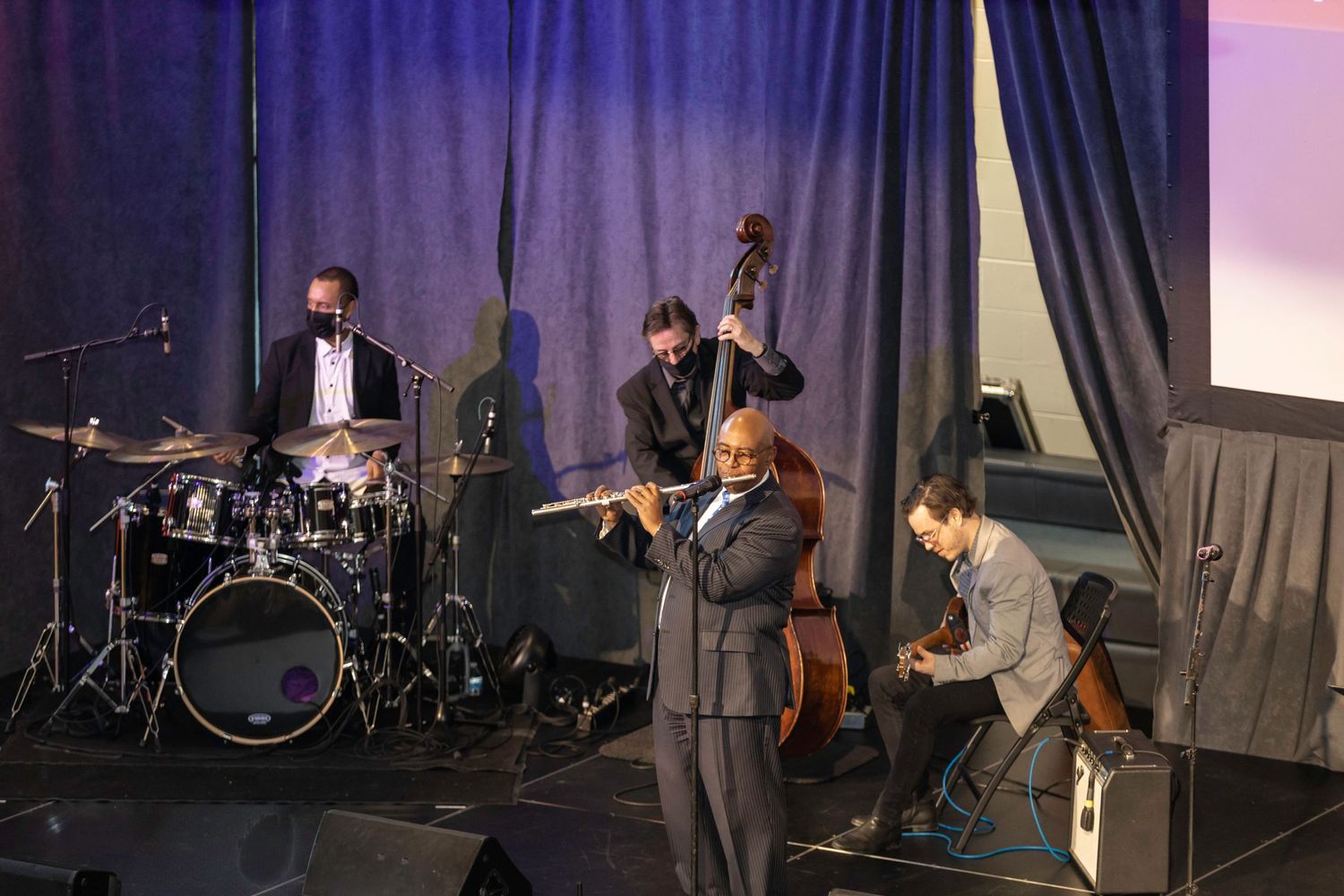 Jazz band performs on stage with drums, bass and other instruments in dim lighting.