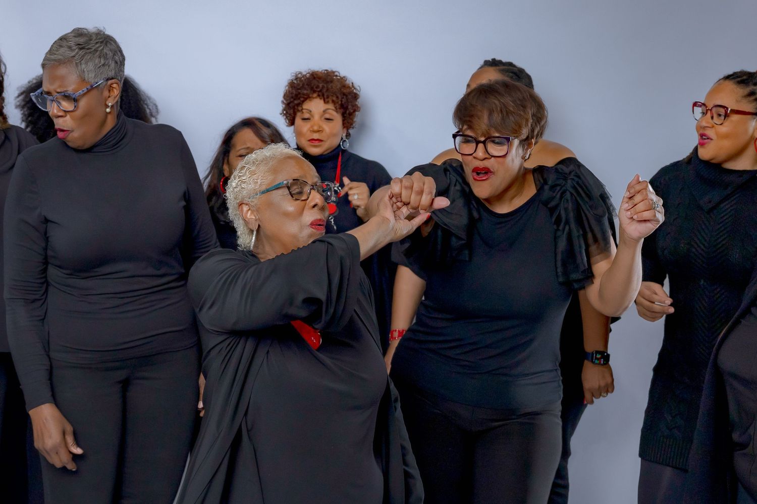 A group of performers in black clothing express dramatic emotions while posing together against a plain background.