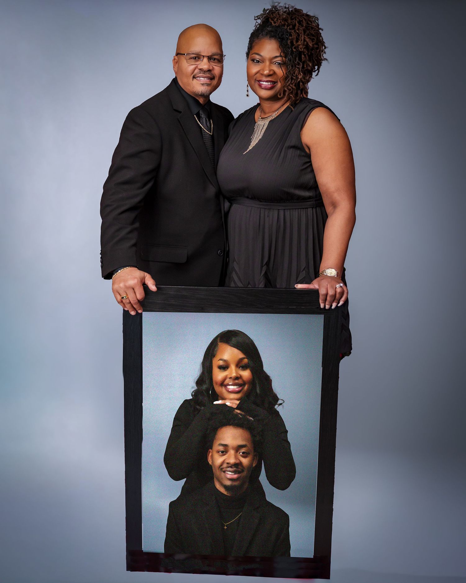 A couple in formal black attire holding a framed photo portrait against a grey background.
