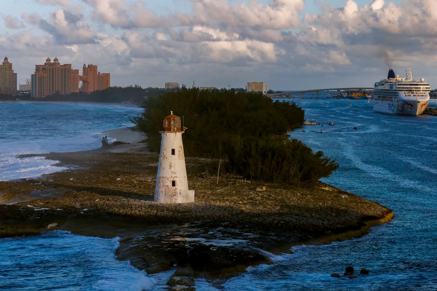 A white lighthouse stands on a rocky outcrop in Nassau Harbor with Atlantis resort and a cruise ship visible in the background.