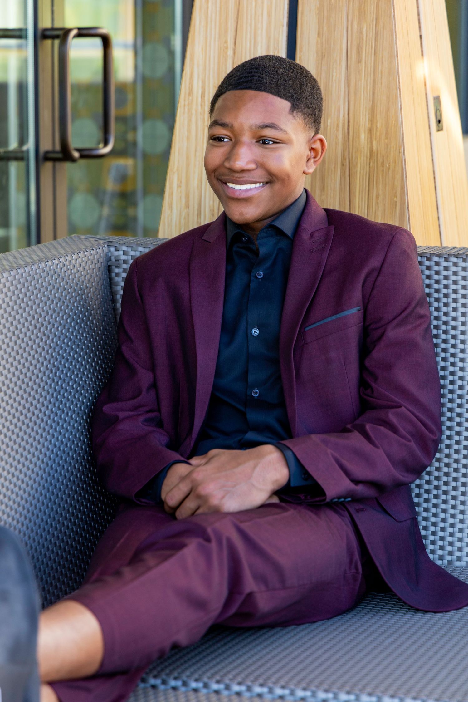 Young man wearing a burgundy suit sits on a grey couch in a modern interior setting.