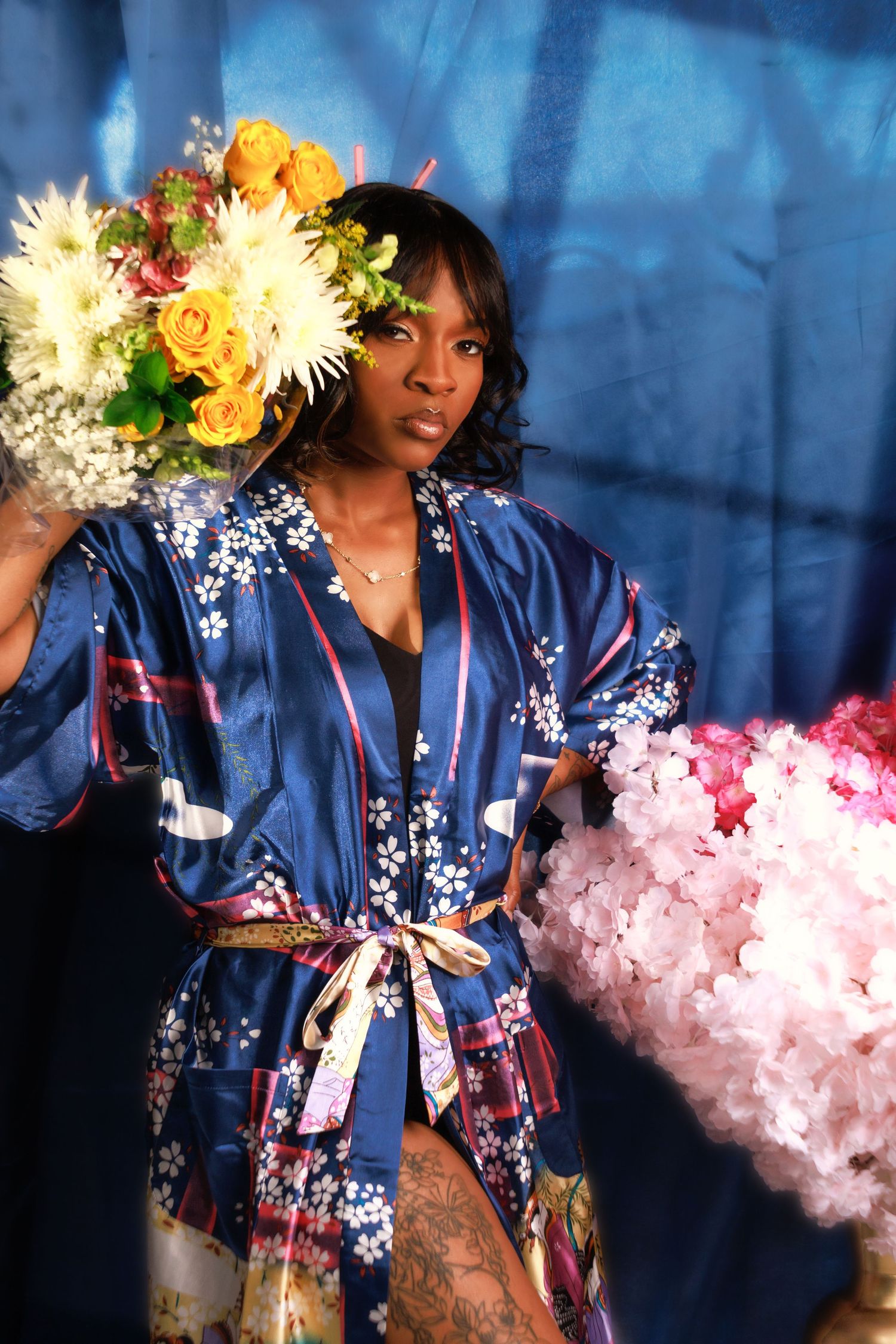 Person wearing a blue floral robe poses with yellow and white flowers against a blue backdrop.