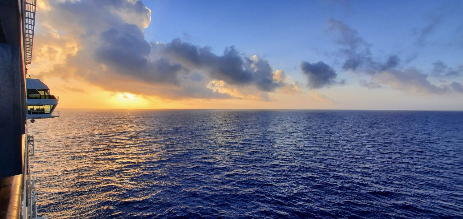 Panoramic sunset view of the ocean horizon with scattered clouds casting golden reflections on deep blue waters.
