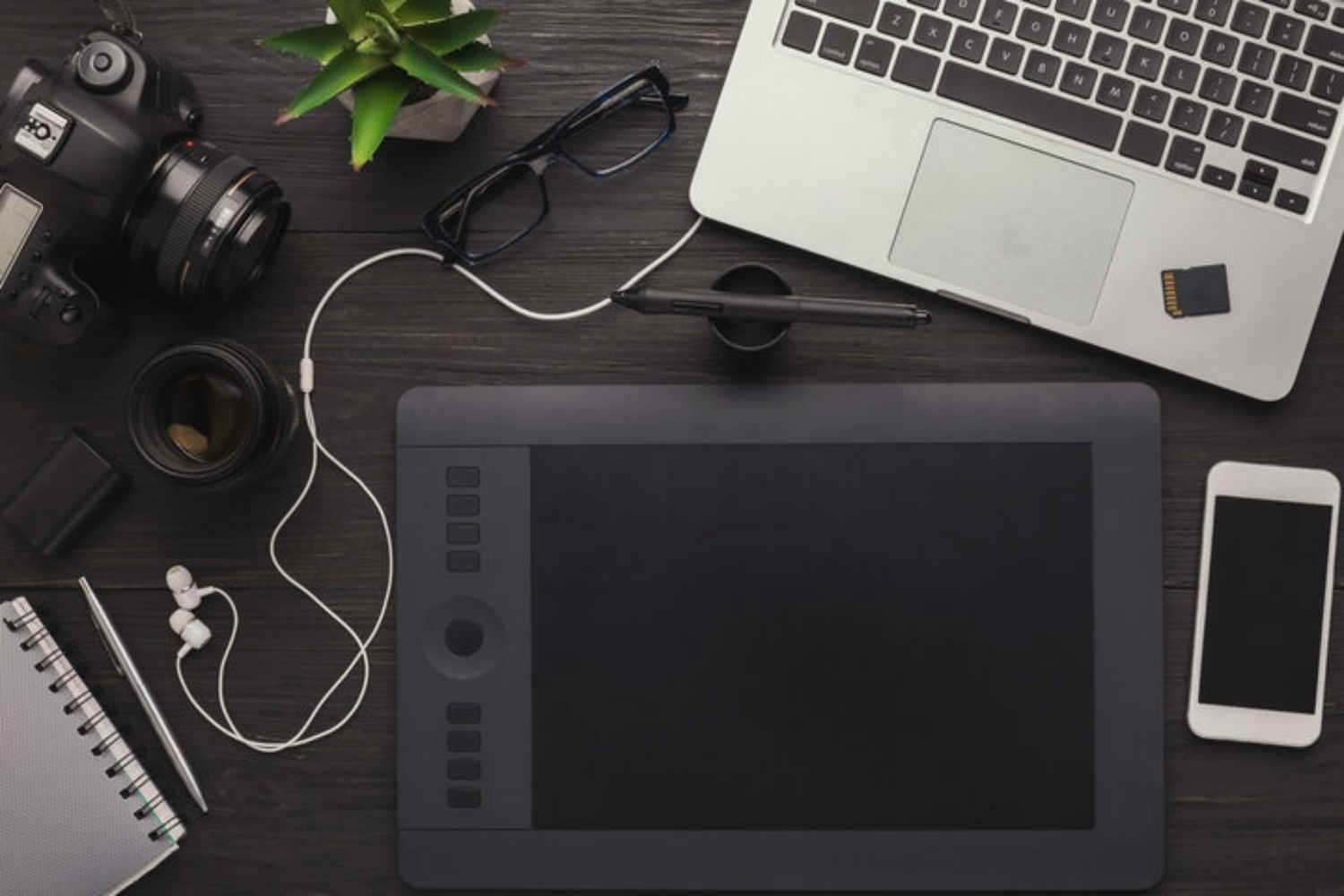 Modern creative workspace with laptop, graphics tablet, camera and smartphone on dark wooden desk viewed from above.
