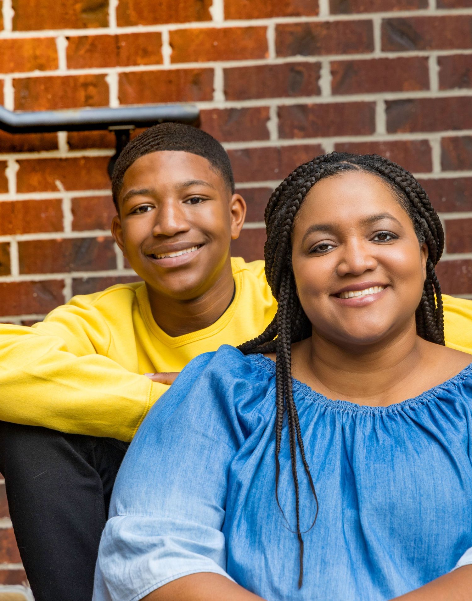Mother and son in yellow and blue shirts smile warmly while sitting against a brick wall background.