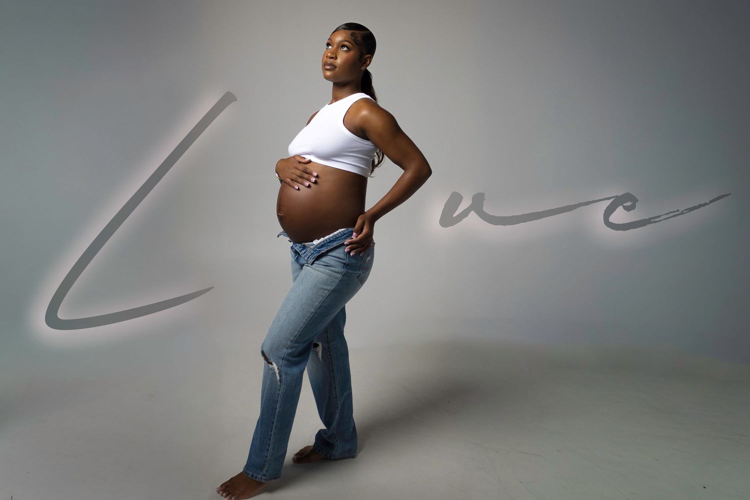 A pregnant person in a white crop top and jeans poses against a gray backdrop with the word 'Love' written in script.