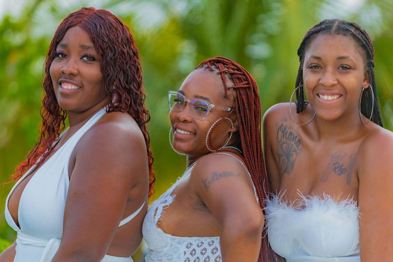 Three women in white outfits smile together against a lush tropical background.