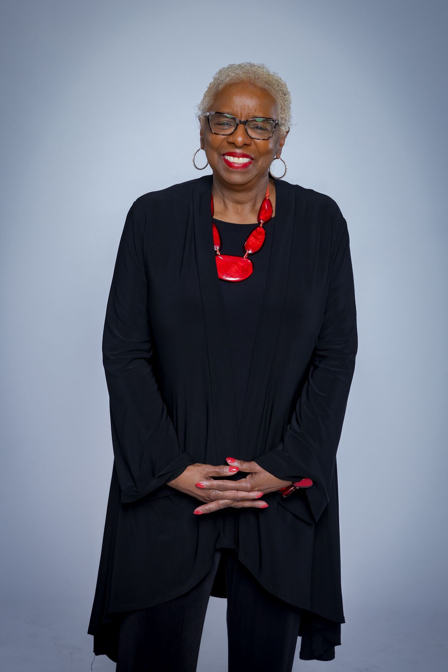 Person in black outfit with red necklace poses against gray background.