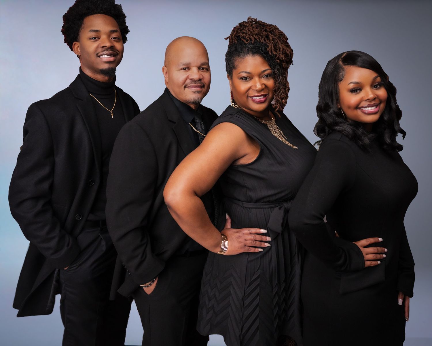 A stylish family posed together in formal black attire against a gray backdrop.