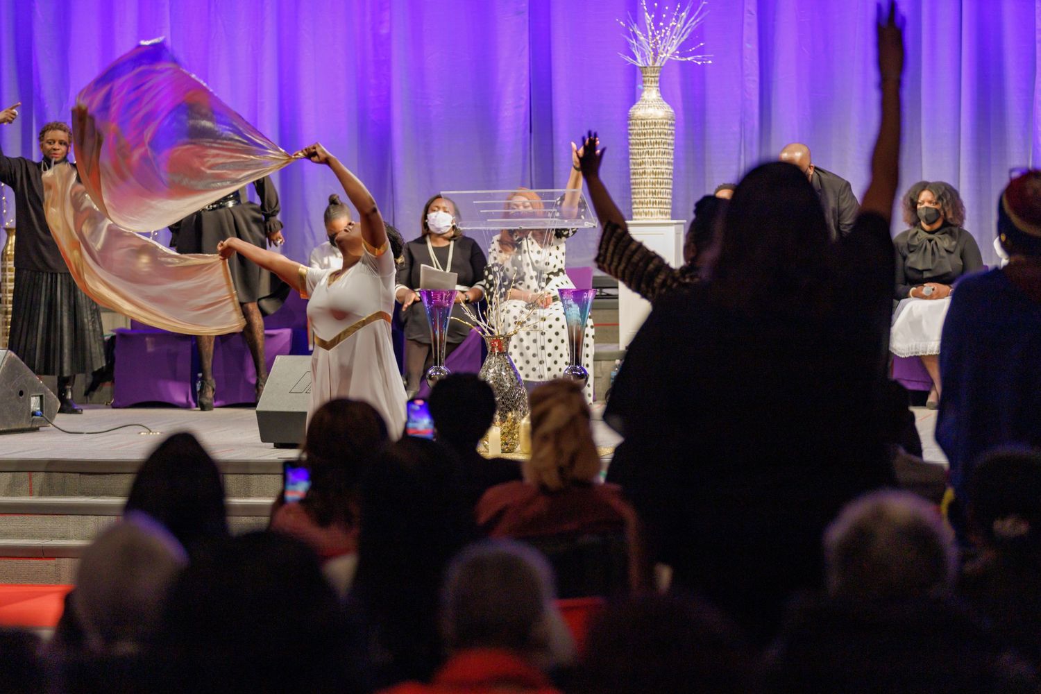 A crowd cheers at a stage performance with purple lighting and decorative elements in the background.