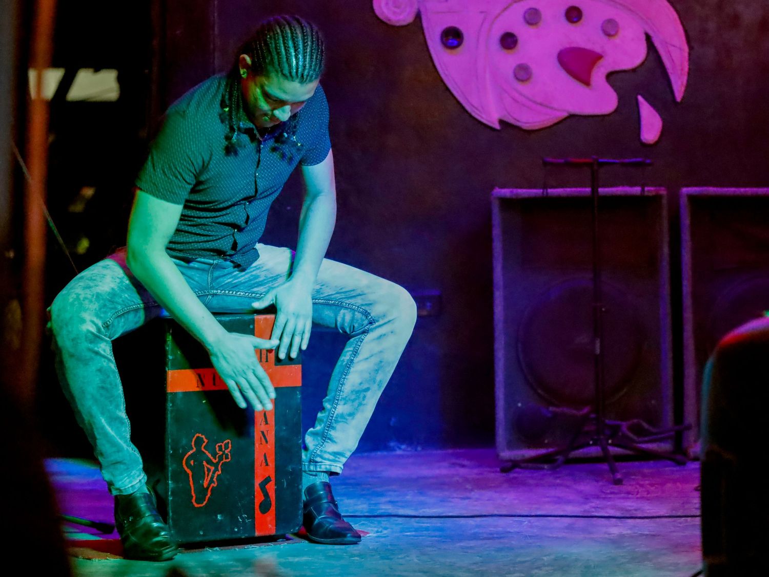 A performer sits on a cajon drum on stage under purple and teal lighting during a live performance.