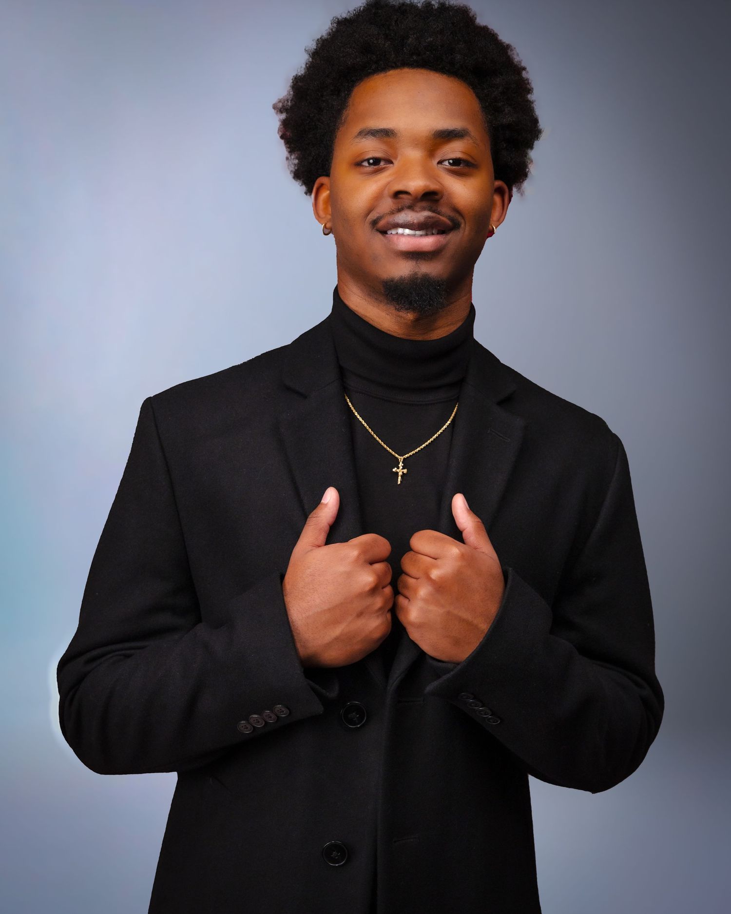 Person in black suit and turtleneck smiles while holding lapels.