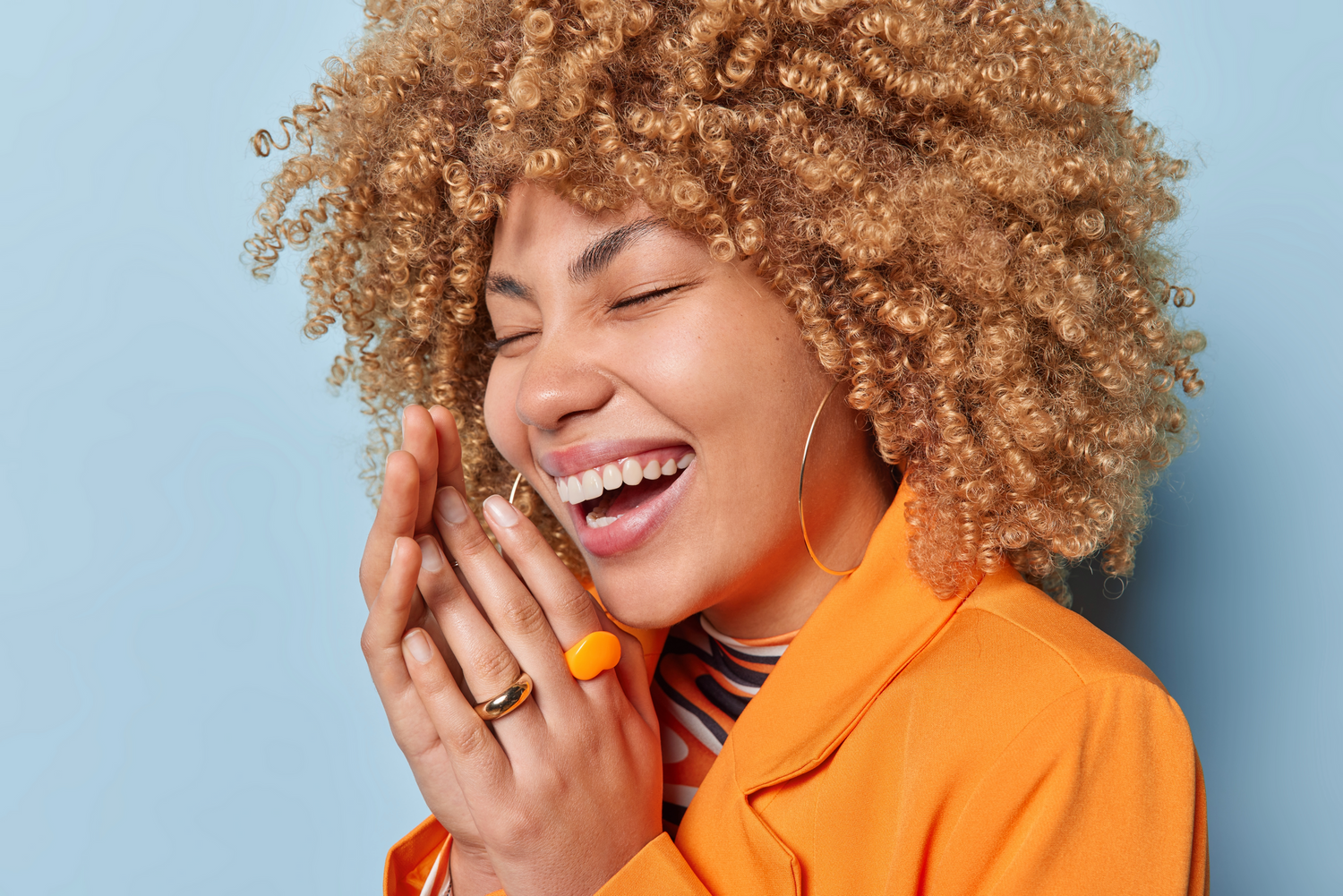 Joyful person with curly blonde hair wearing orange jacket laughs with eyes closed against blue background.