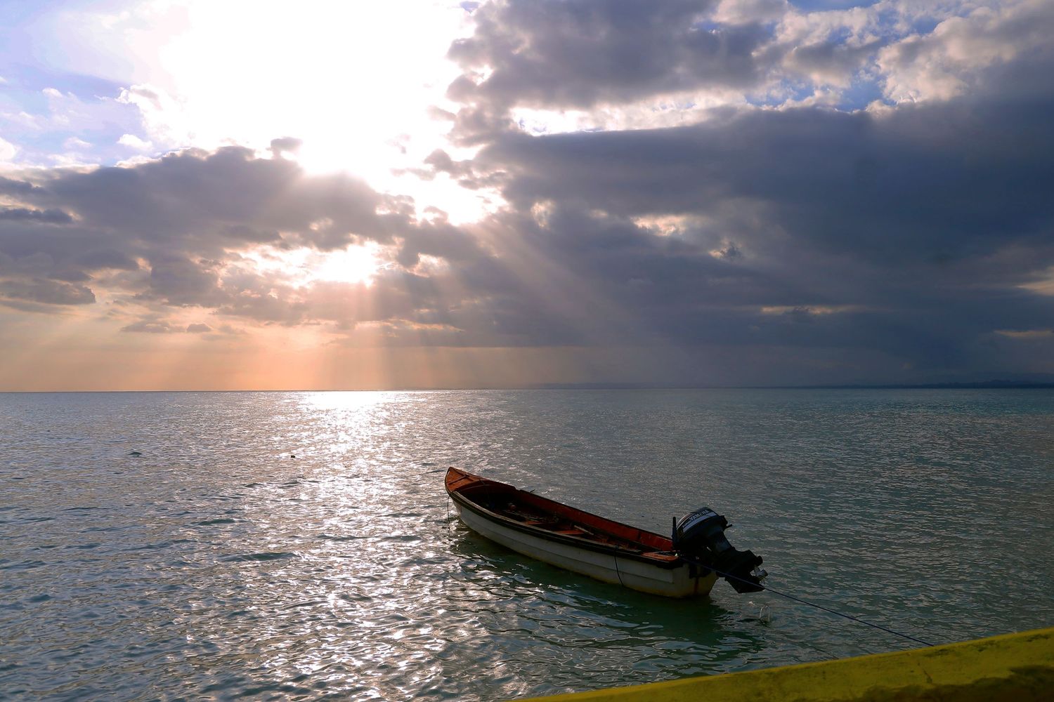 Small boat floating on calm ocean waters at sunset with dramatic sunbeams piercing through cloudy sky.