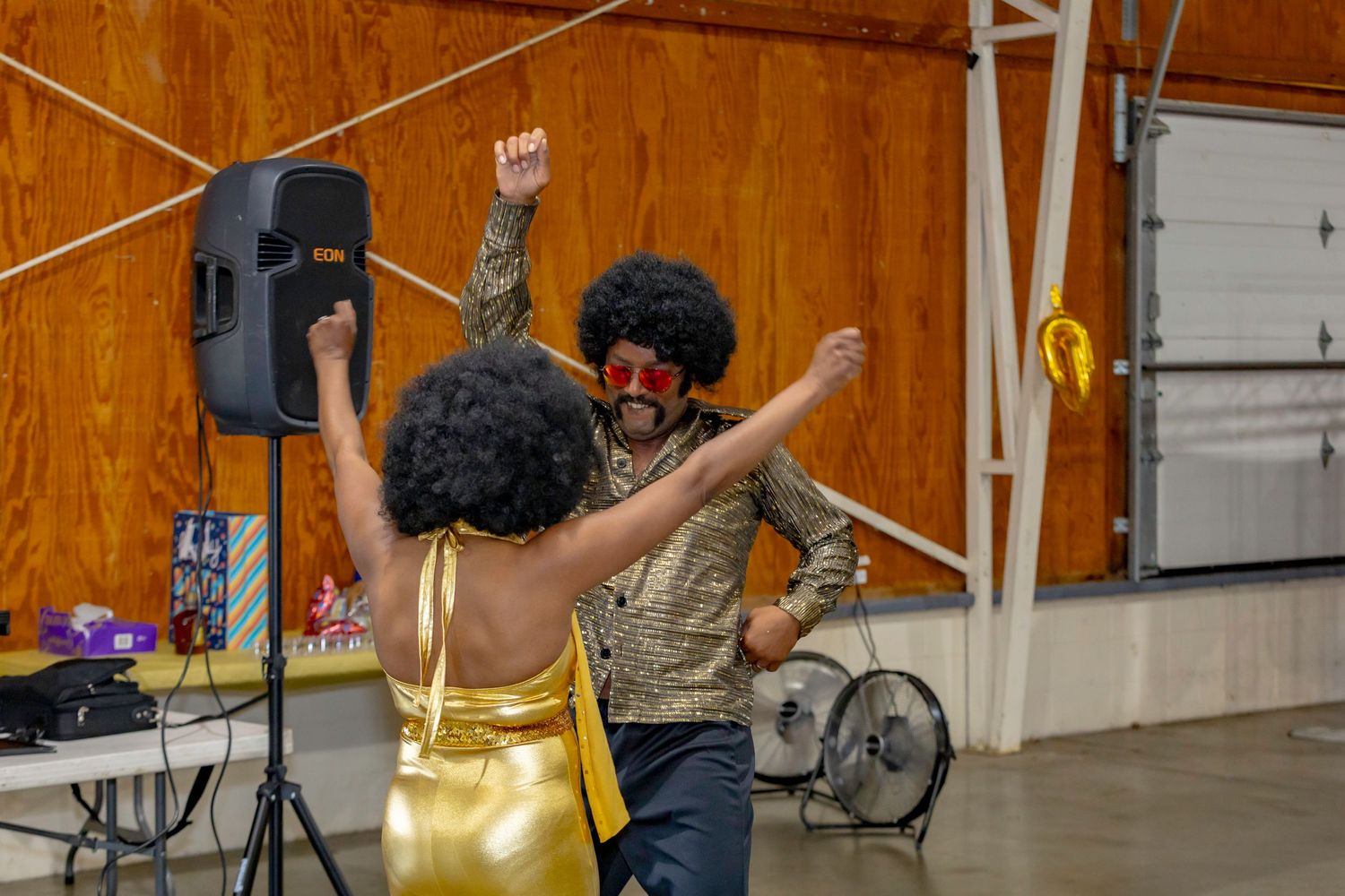 Two dancers in festive attire move energetically to music in front of a speaker system.