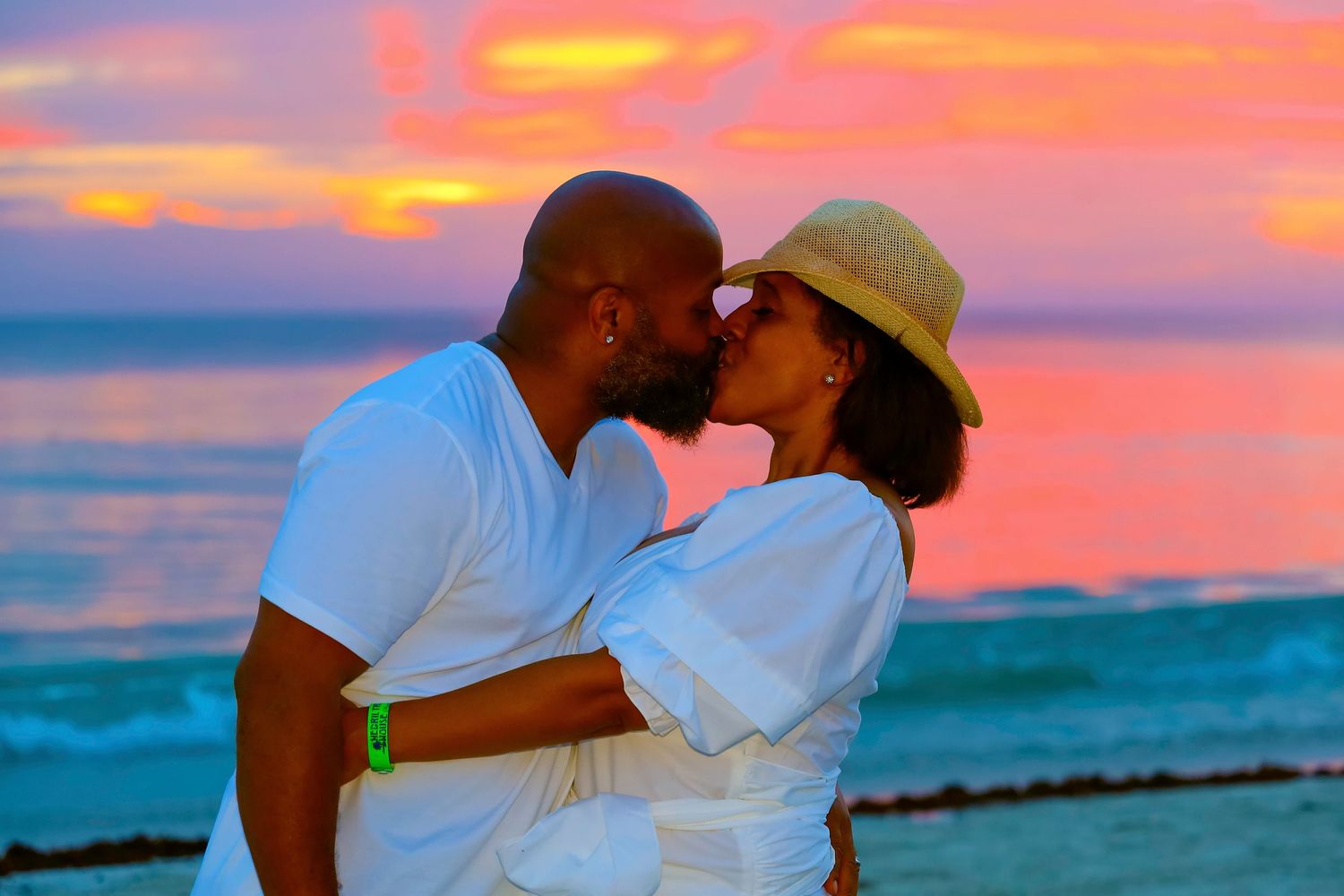 A romantic couple shares a kiss on the beach at sunset with vibrant orange sky and turquoise waters in the backdrop.
