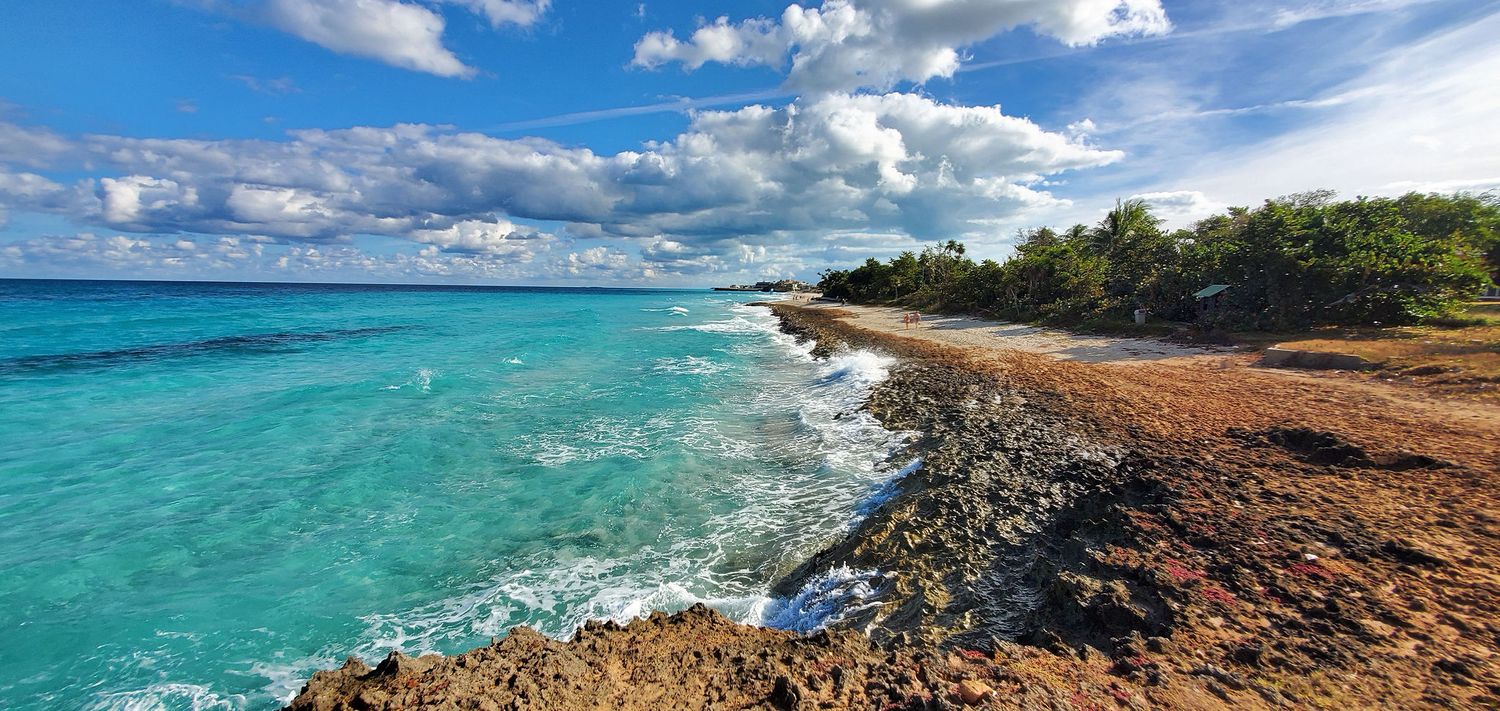 Turquoise waters crash against a rocky shoreline under puffy white clouds at a tropical Caribbean coastline.