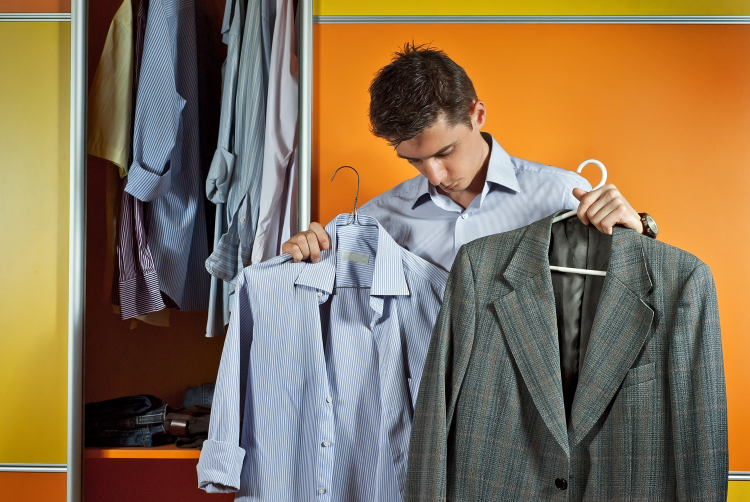 Person choosing between a blue dress shirt and gray suit jacket while standing at an open closet with orange walls.