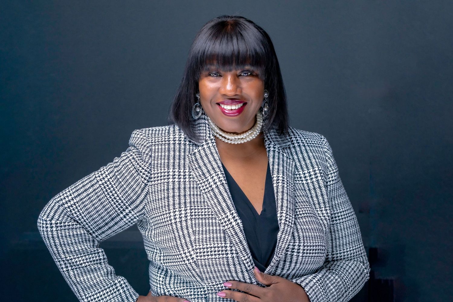 Professional headshot of a person wearing a plaid blazer with a black top and pearl necklace against dark background.