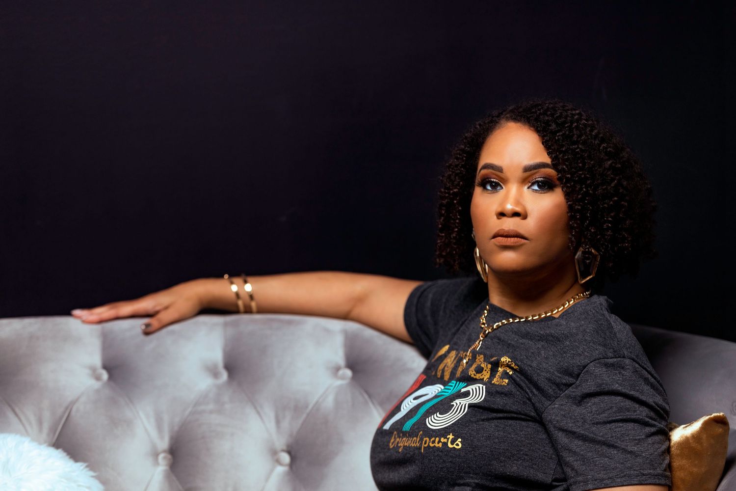 Woman in casual black t-shirt poses on tufted white leather couch against dark backdrop.