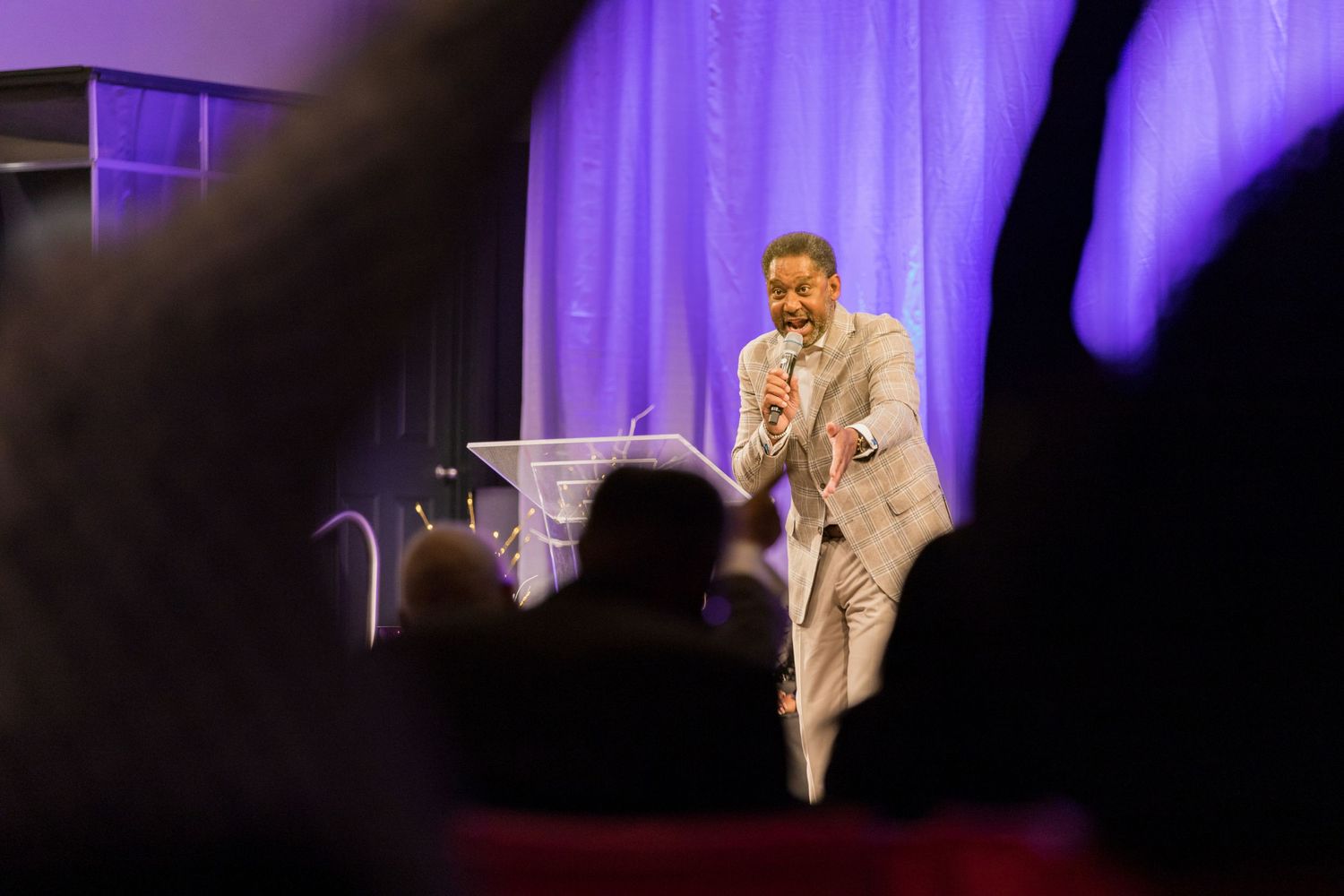 A speaker presenting at a podium on stage with purple lighting and curtains.