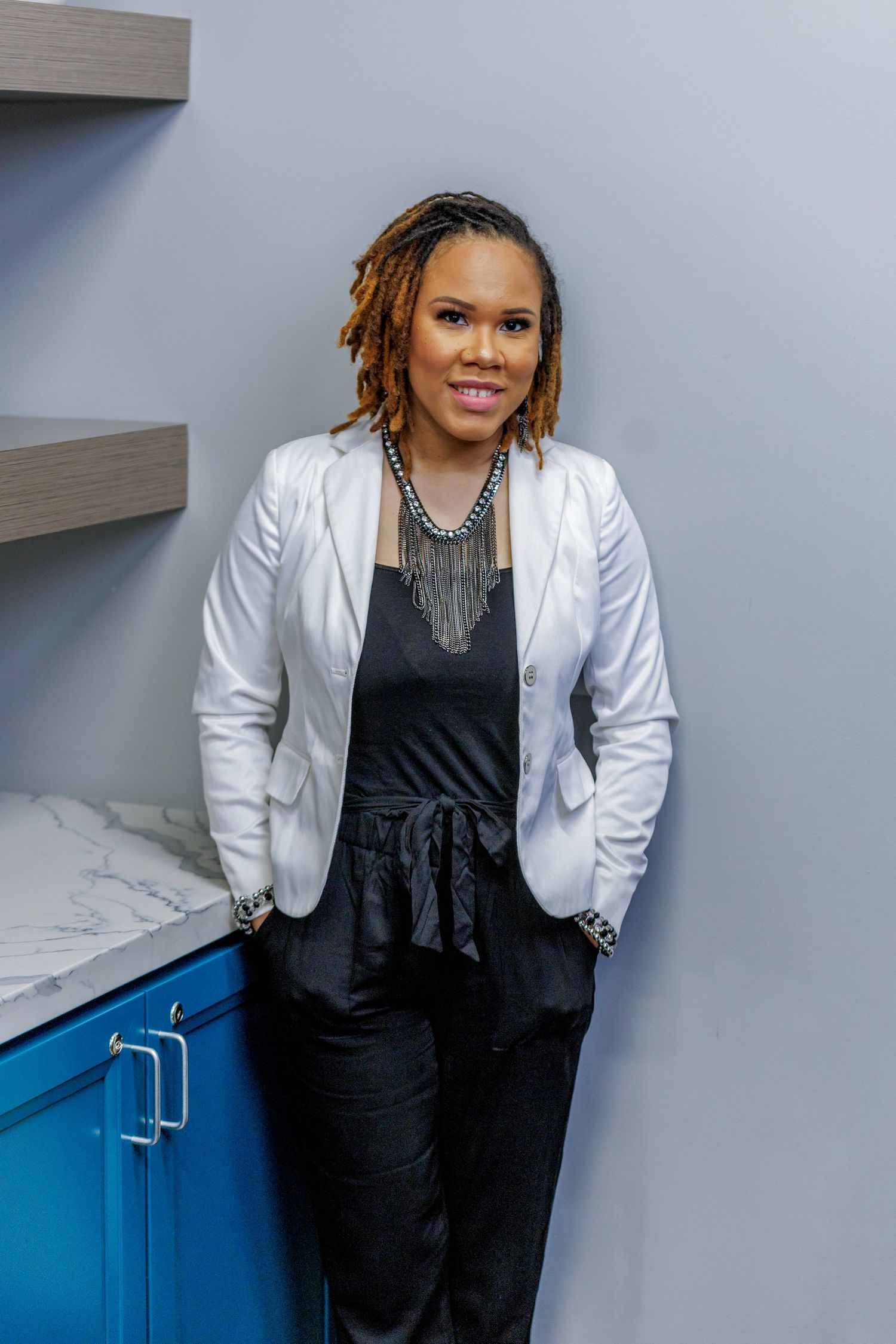 Professional portrait of a person in a white blazer and black outfit with statement necklace leaning against blue cabinets.
