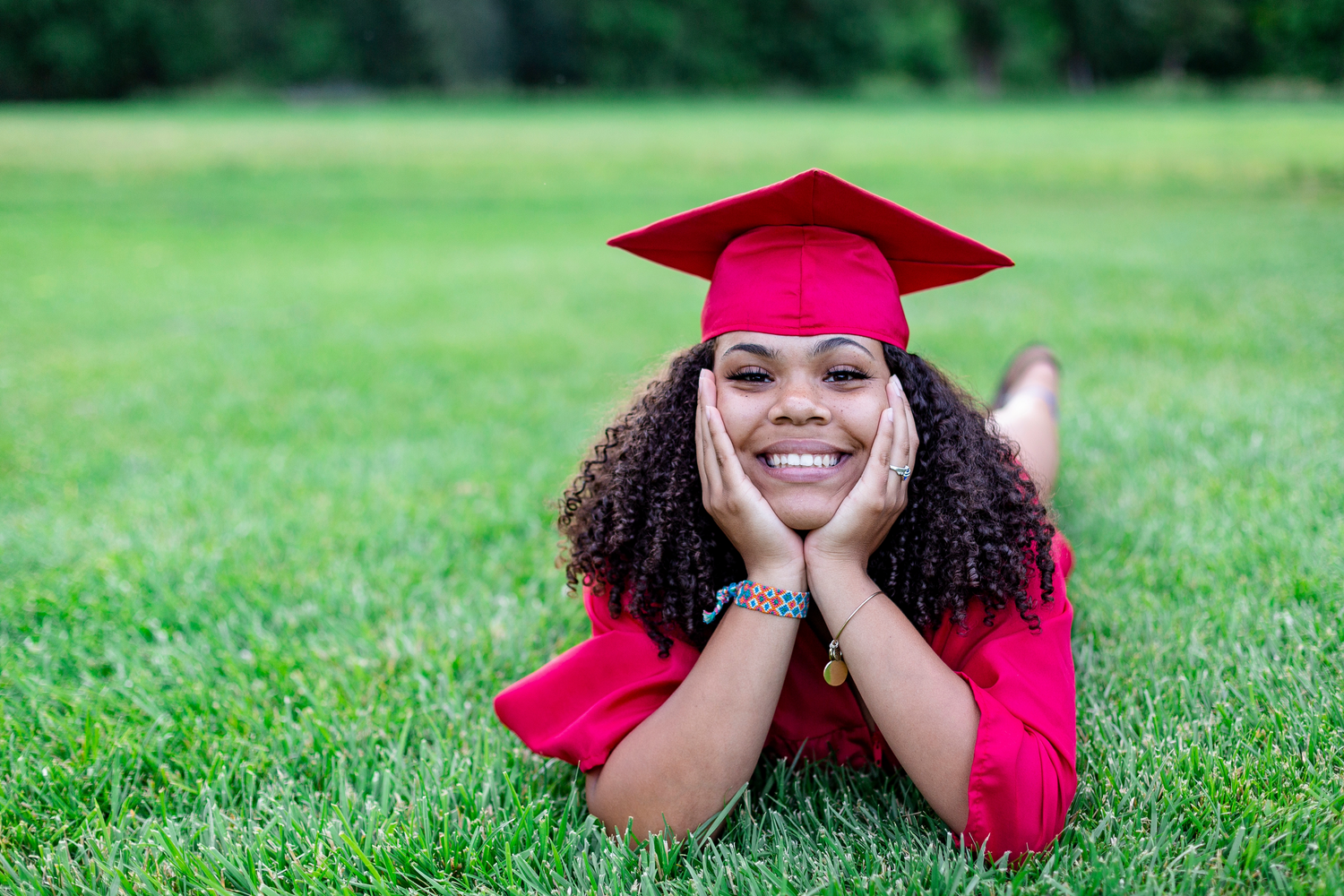 Graduate in red cap and gown lies on grass during outdoor graduation photo session.