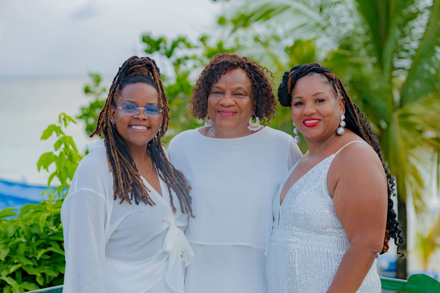 Three women in white dresses pose together at a tropical outdoor location.