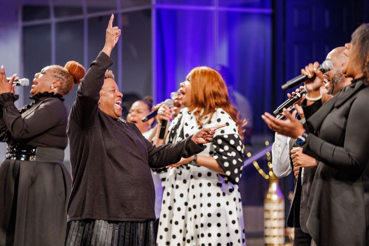 Gospel choir members raise their hands in worship while singing in polka dot and black attire.