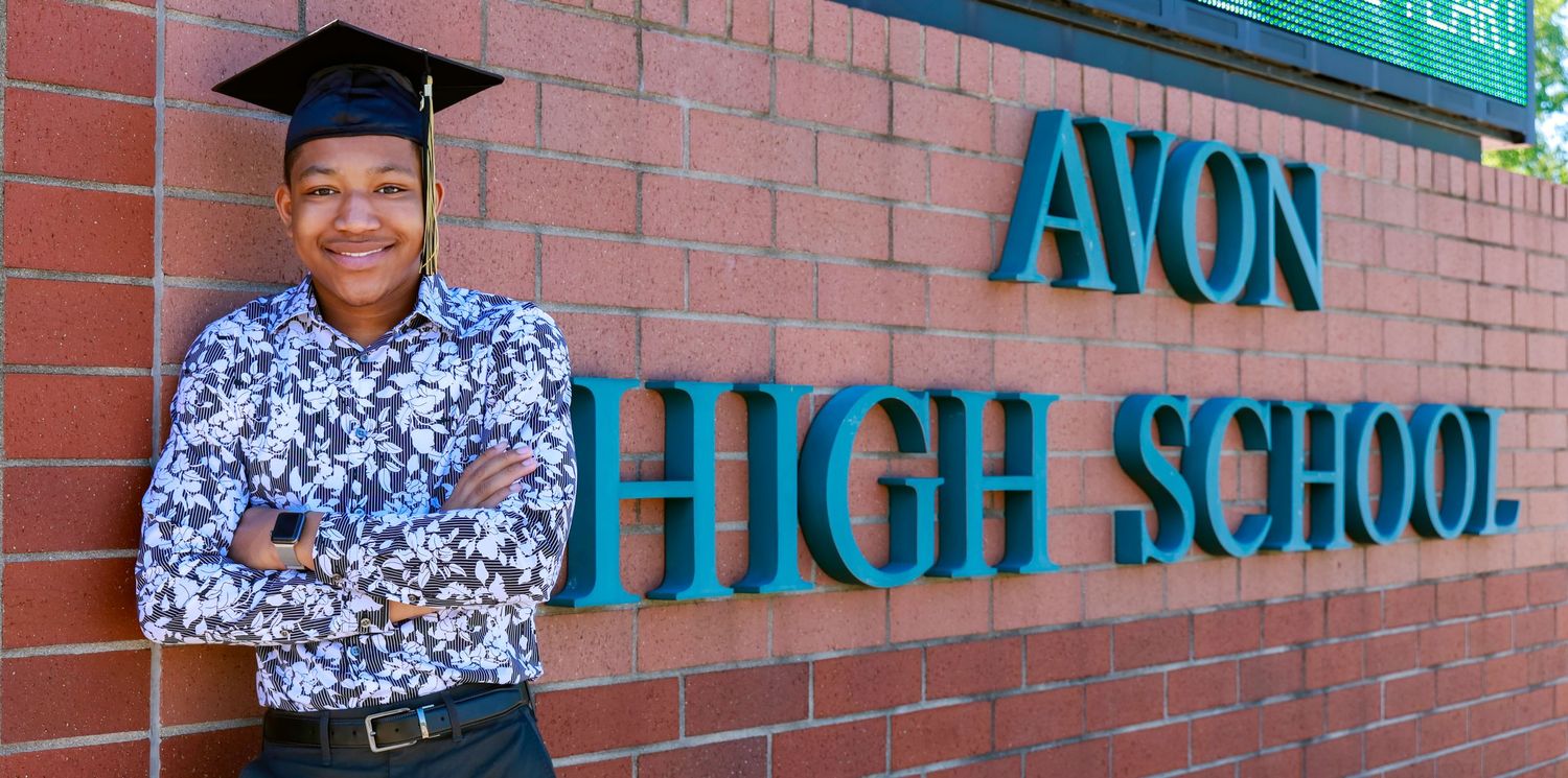 A graduate poses proudly in front of Avon High School brick wall signage.