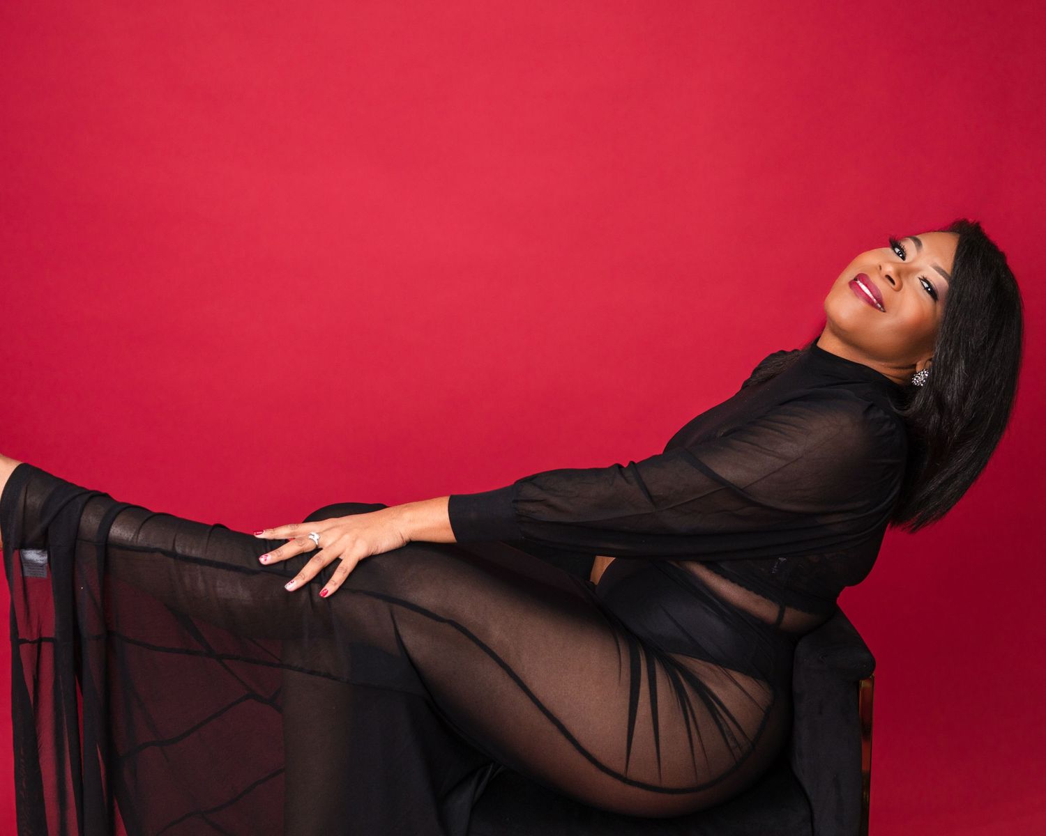 A figure in a black sheer dress poses expressively against a vibrant red backdrop.