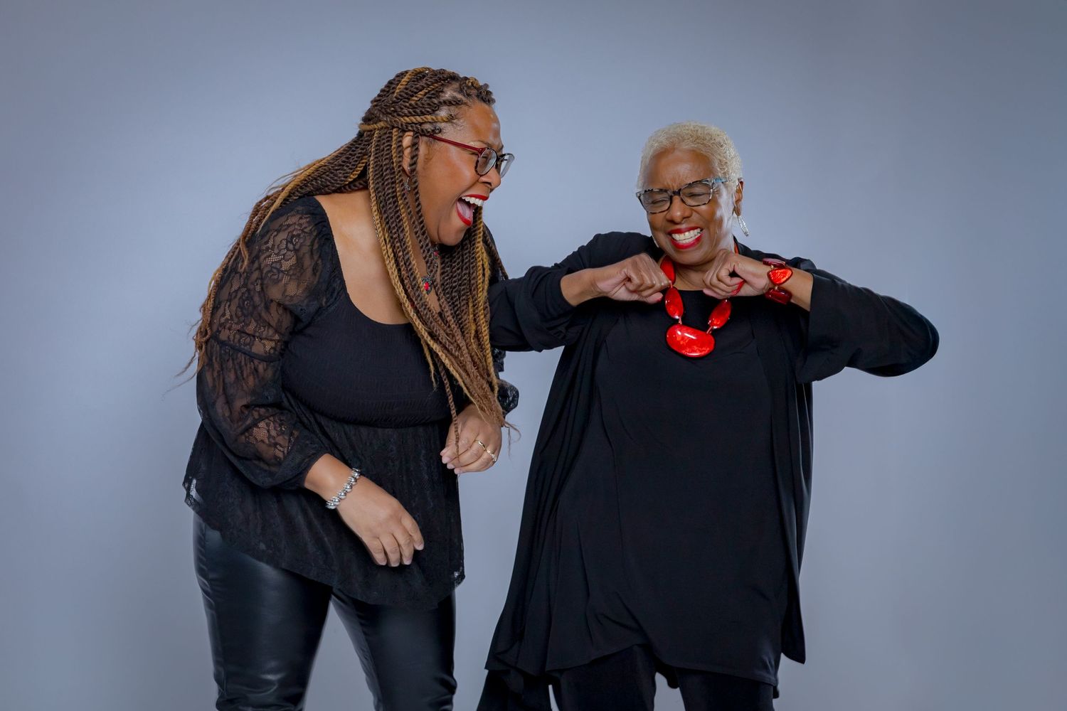 Two people wearing black outfits share a joyful moment while laughing and gesturing against a gray background.