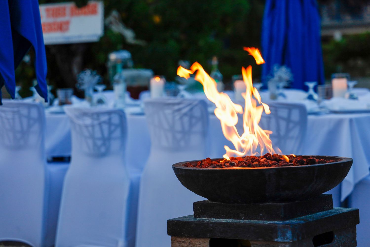 Fire burns in a decorative bowl against elegant outdoor table settings with blue decor and glassware in the background.