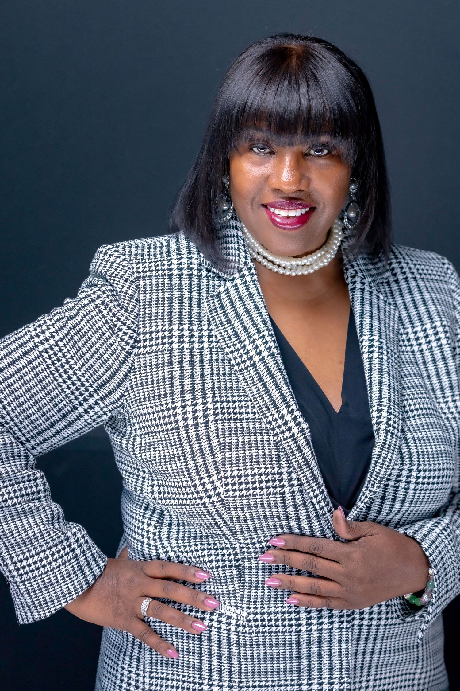 Professional portrait in black and white plaid blazer with pearl necklace against dark background.