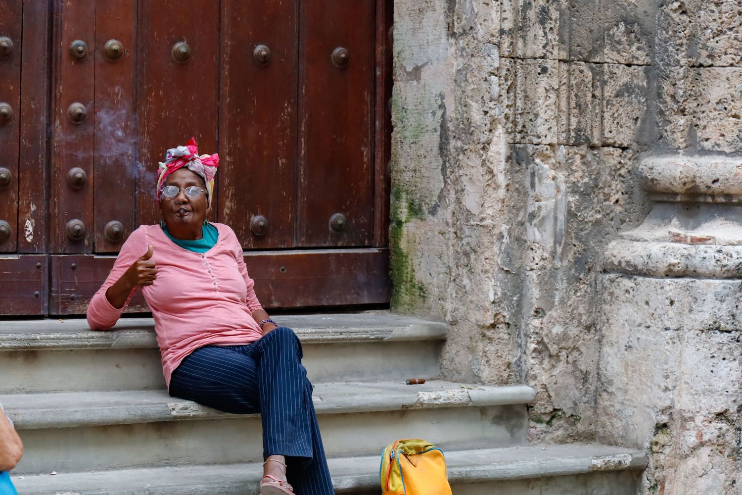 A local Cuban woman in a pink shirt and headscarf smoking a cigar sits on stone steps near a wooden door.