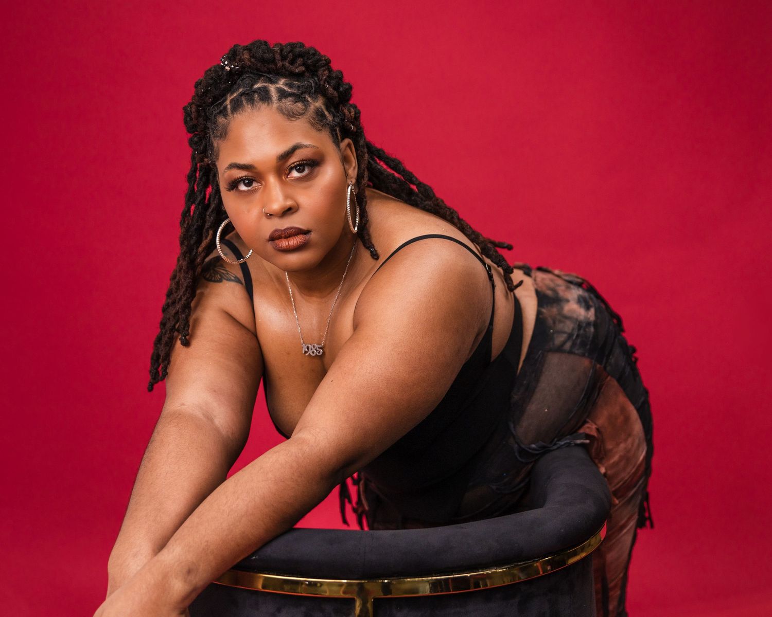 A glamour portrait against a red backdrop featuring a model in black attire posing on a chair.