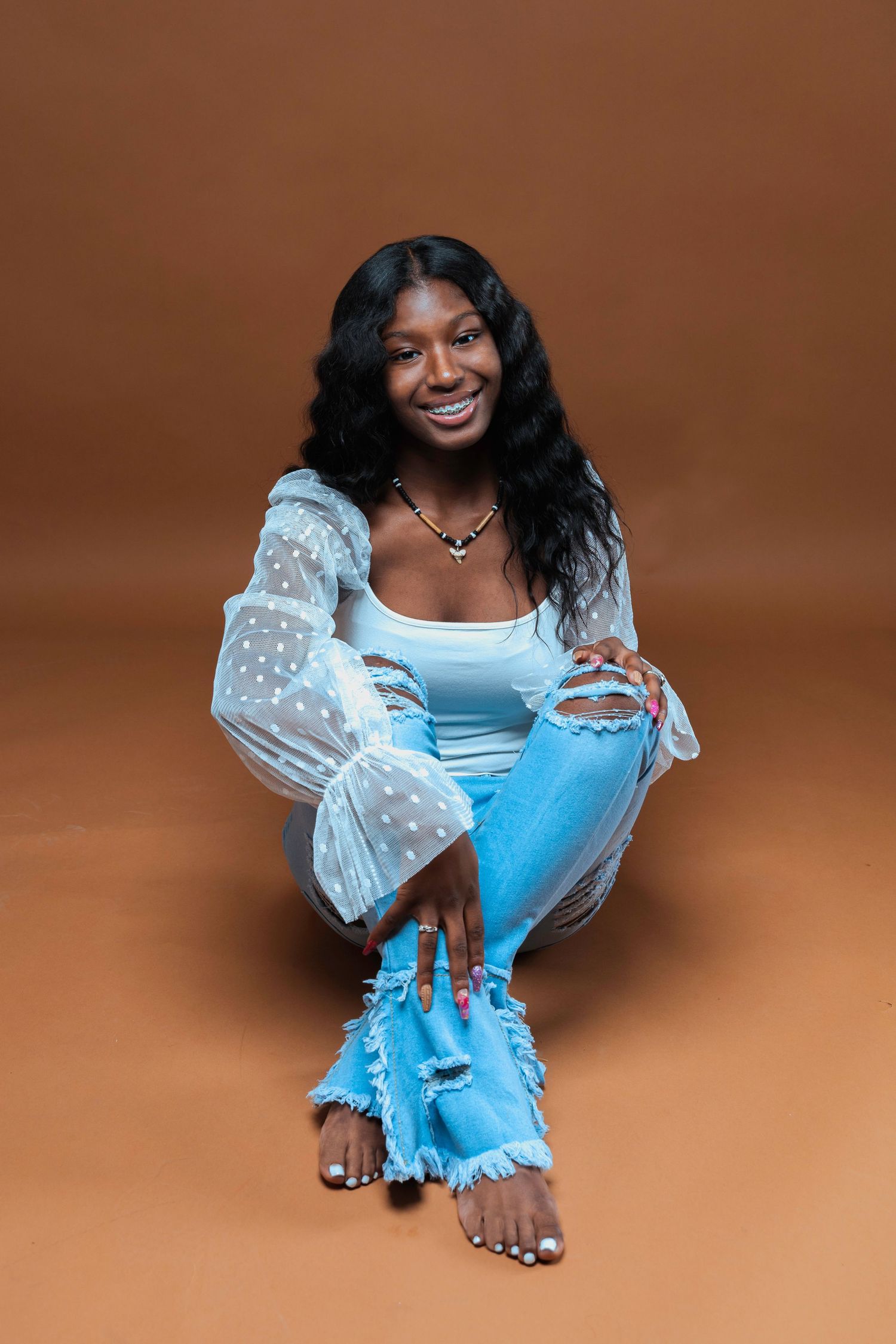 Woman in white lace top and distressed jeans sitting cross-legged on brown backdrop during fashion photoshoot.