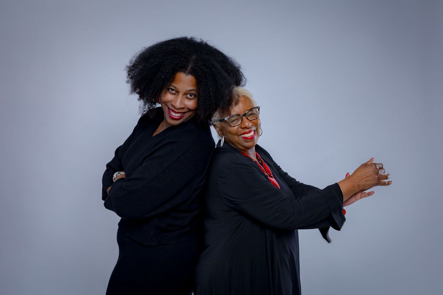 Two business professionals in black attire pose back-to-back with confident smiles against a gray backdrop.