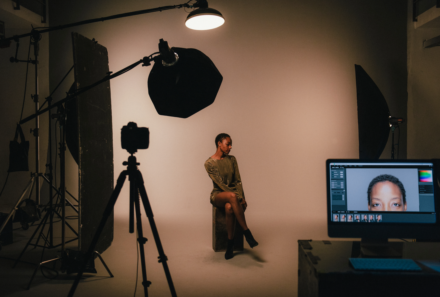 Professional photography studio setup with lighting equipment, camera on tripod, backdrop, and monitor display for photo session.