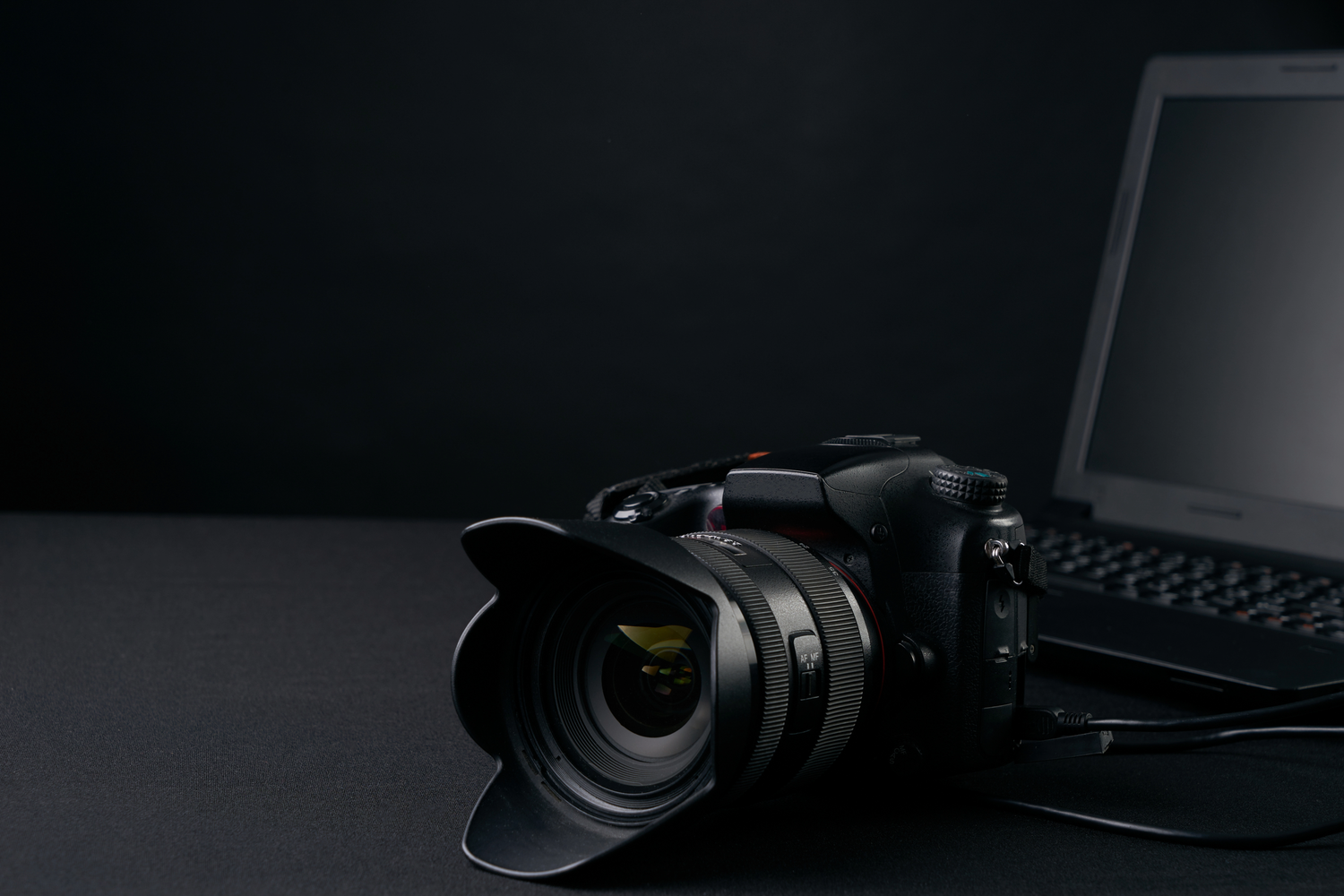 A professional black DSLR camera with lens hood sits on dark surface next to laptop keyboard in low-key lighting.