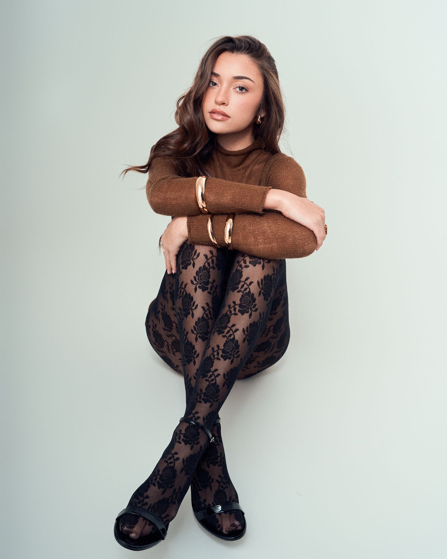 Model in brown turtleneck and lace tights sitting in elegant posed position.