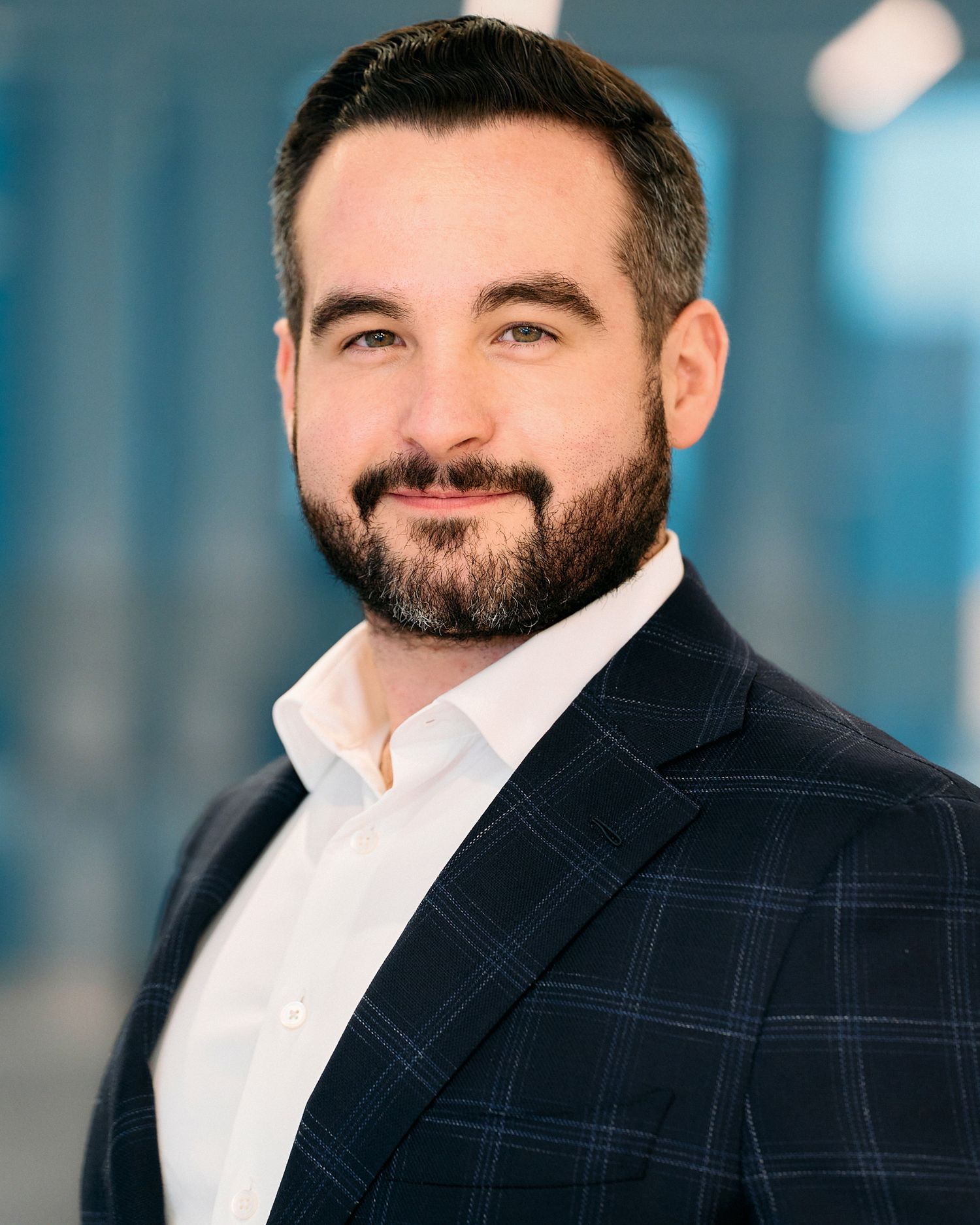 Professional headshot portrait of person in navy suit jacket and white dress shirt against blurred blue background.