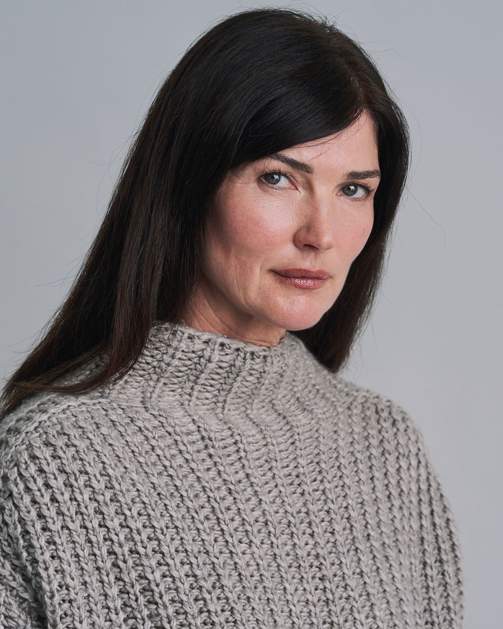 A woman with long dark hair wearing a gray knit turtleneck sweater poses against a light gray background.