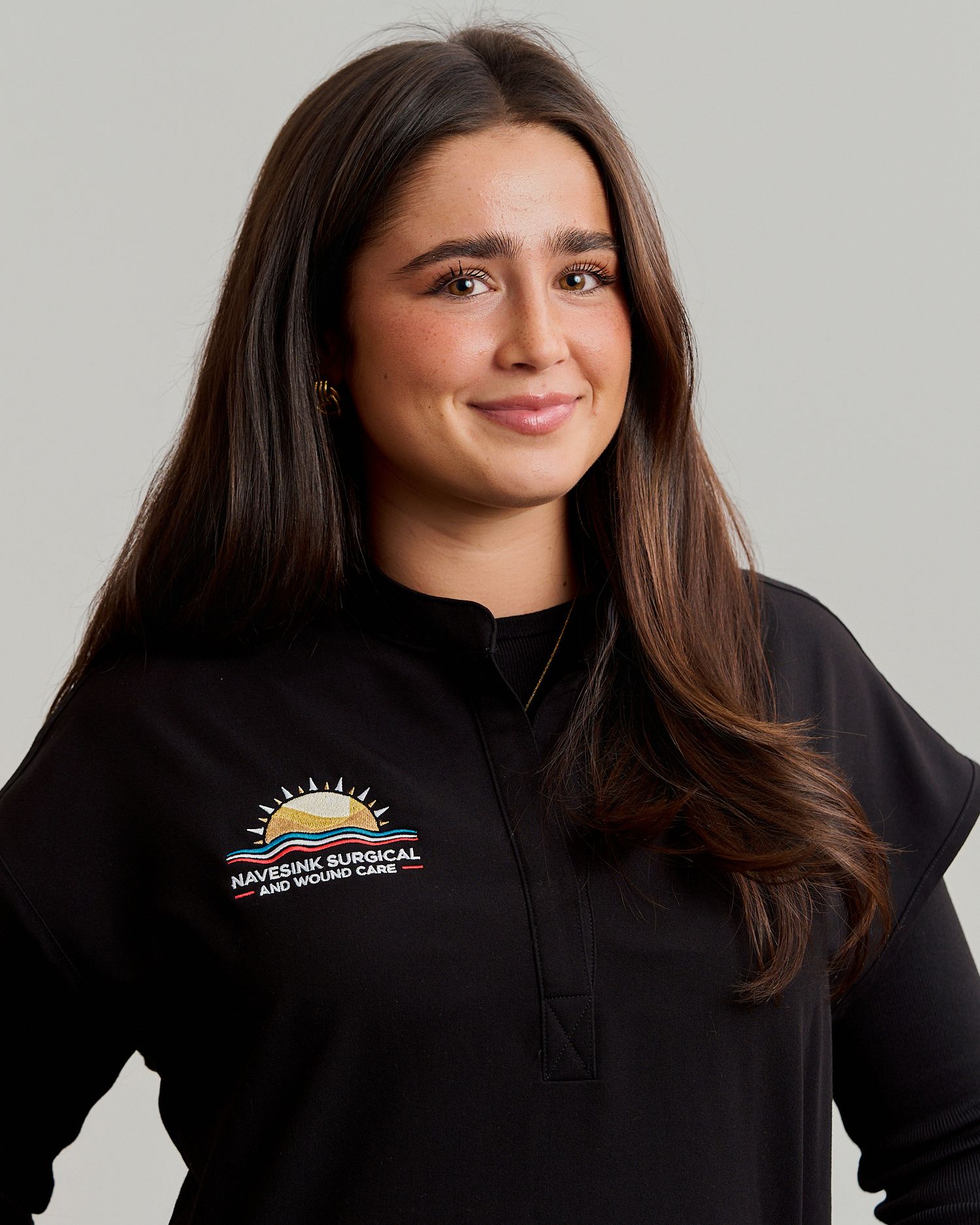 Woman in black Navesink Surgical and Wound Care branded quarter-zip jacket smiling against gray background.