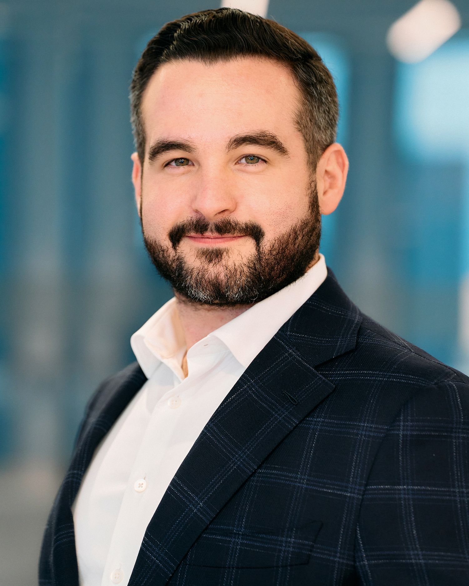 Professional headshot of person in navy plaid blazer and white shirt against blurred office background.