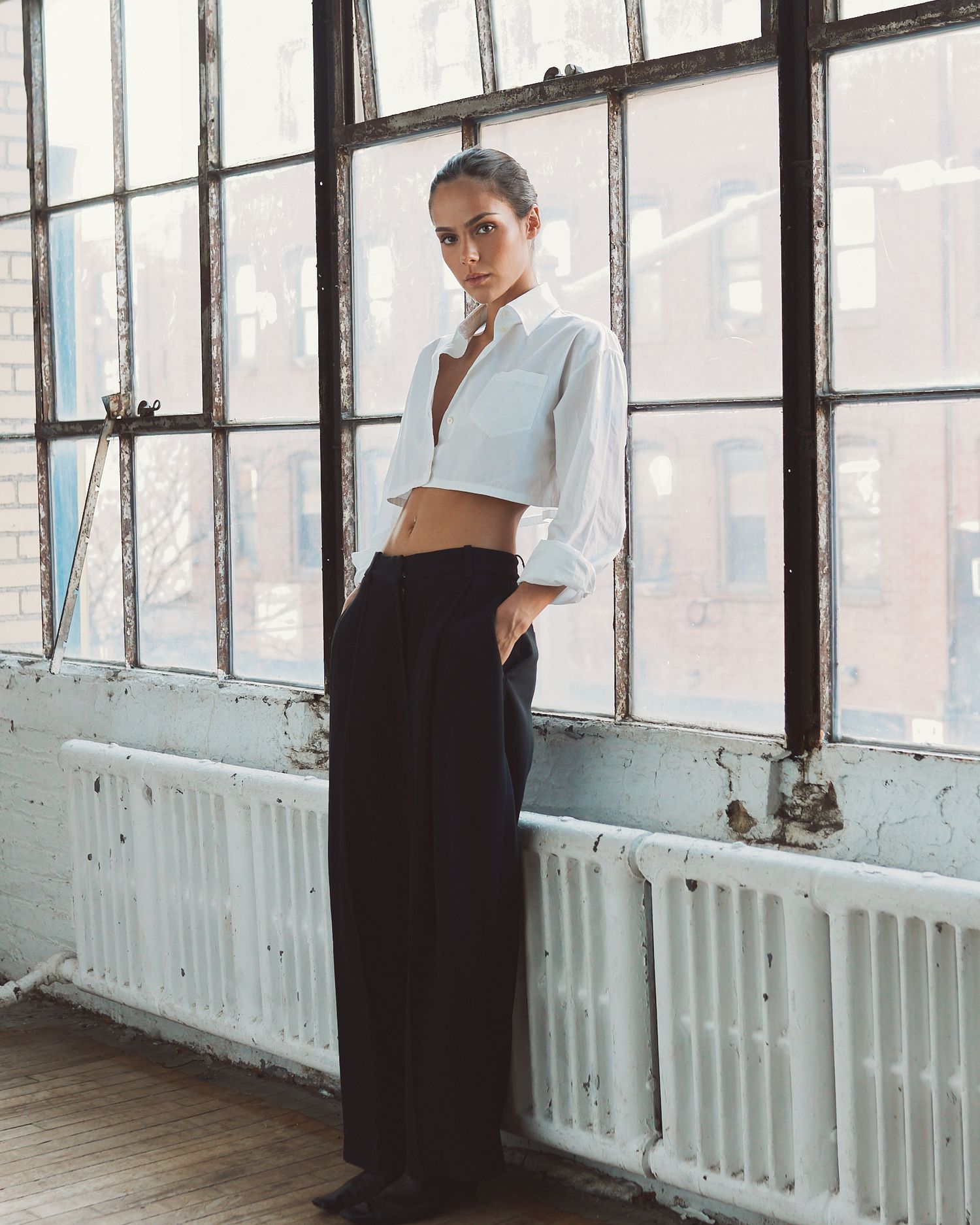 Figure in a white top and black skirt stands near large industrial windows in a bright sunlit room.
