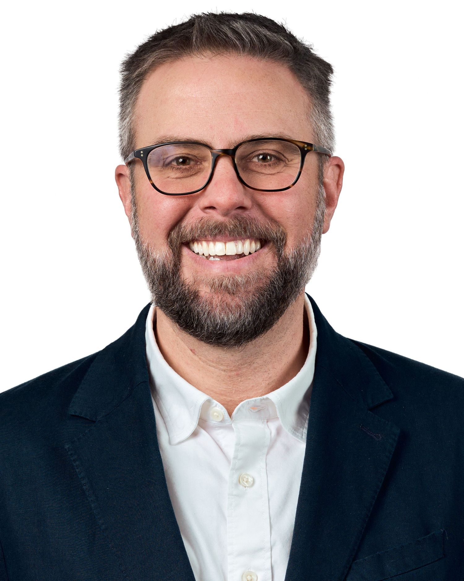 Smiling man with glasses and beard wearing a navy blazer and white shirt against a white background.