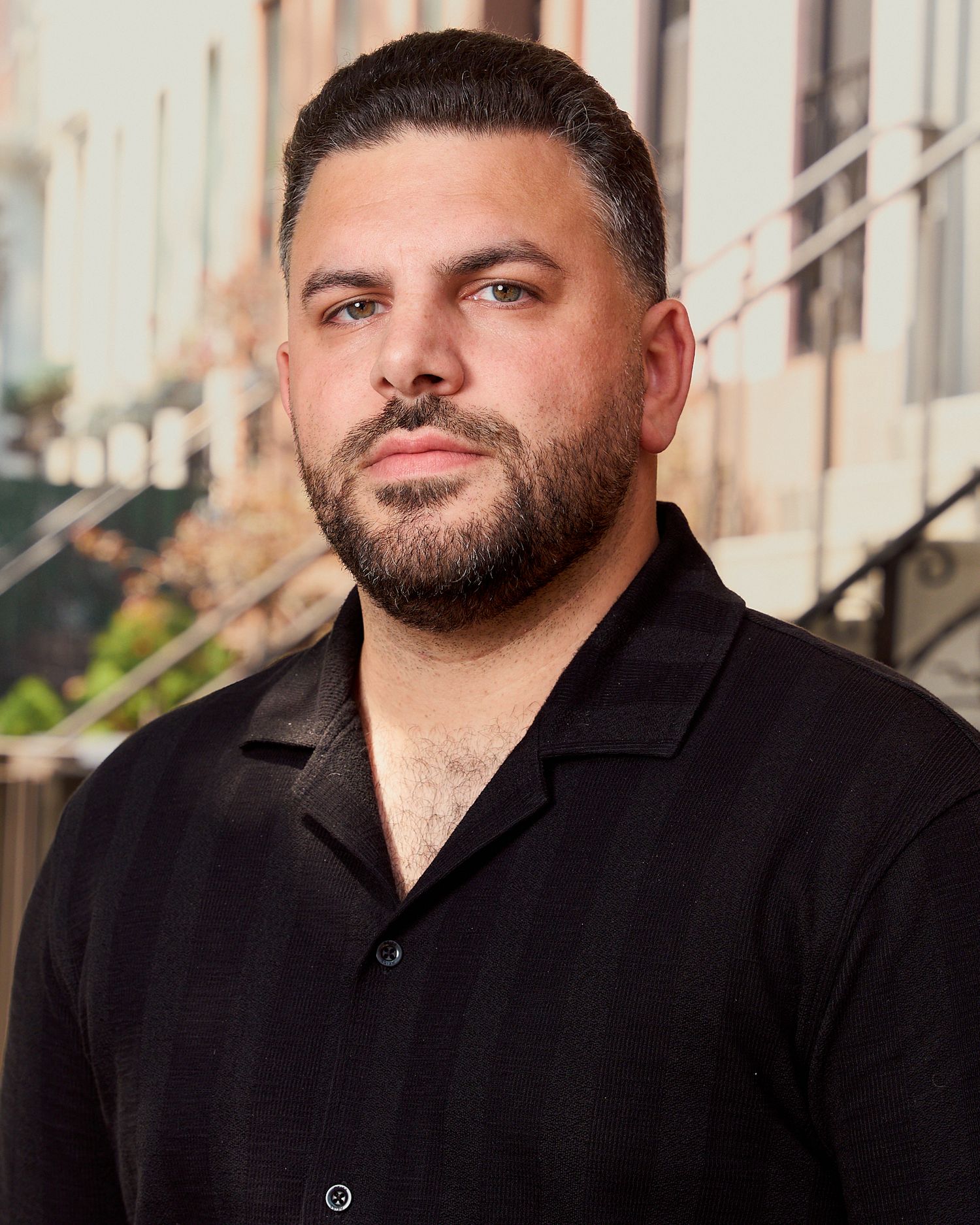 Professional headshot of a bearded man in a black shirt posing outdoors on an urban street.
