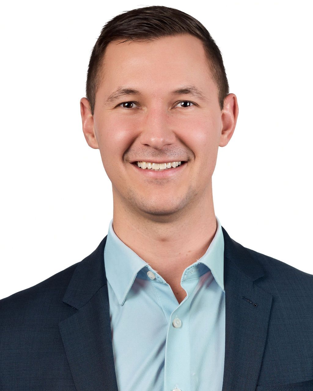 Professional headshot of a smiling young man in a light blue shirt and dark blazer against a white background.