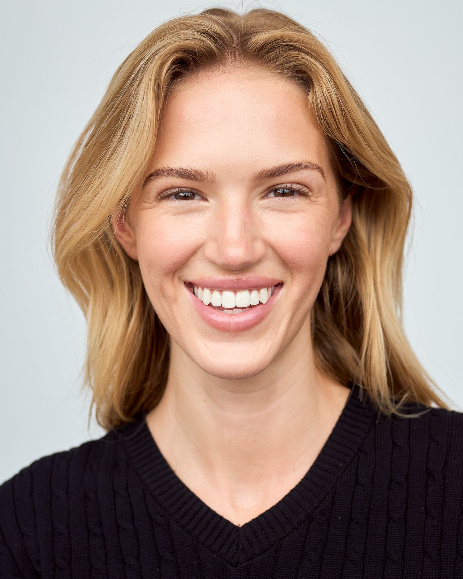 Smiling blonde woman in black V-neck sweater against light gray background, professional headshot photo.